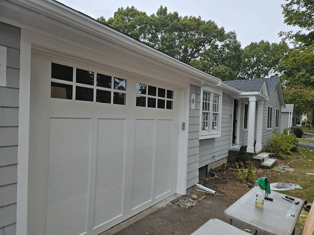A white, multi-paneled garage door with windows, attached to a gray-sided suburban house on a cloudy day.