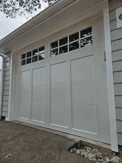 A white, multi-paneled garage door with top window inserts, mounted on a house with light gray shingle siding.