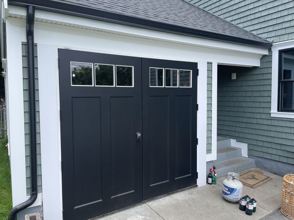 A black carriage-style garage door with windows, attached to a house with dark green shingles and white trim.