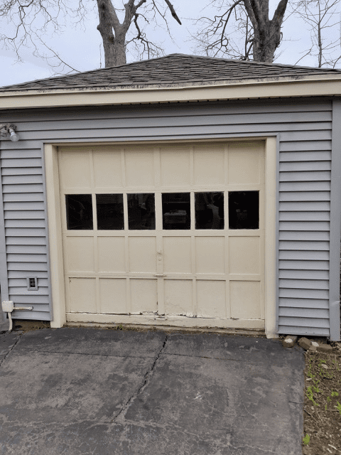 A detached garage with grey vinyl siding, a cream-colored door with six window panes, and a cracked asphalt driveway.