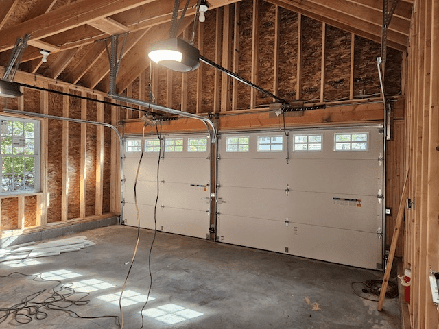 Interior of an unfinished wood-framed garage with two white sectional garage doors and exposed ceiling rafters.