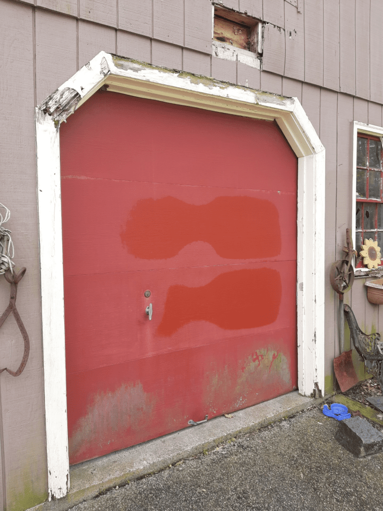 A red garage door with worn white trim in a weathered wooden structure, with patches of paint and signs of age.