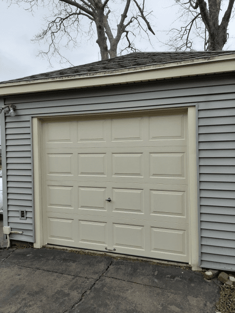 A single-car garage with light tan paneled doors and gray vinyl siding under a dark shingled roof on an overcast day.