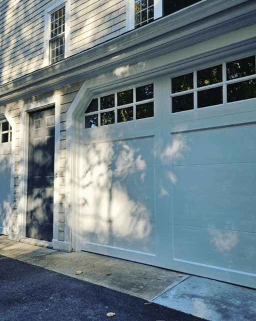 A white garage door with rectangular windows and a side entry door, dappled with light and shadows from nearby trees.