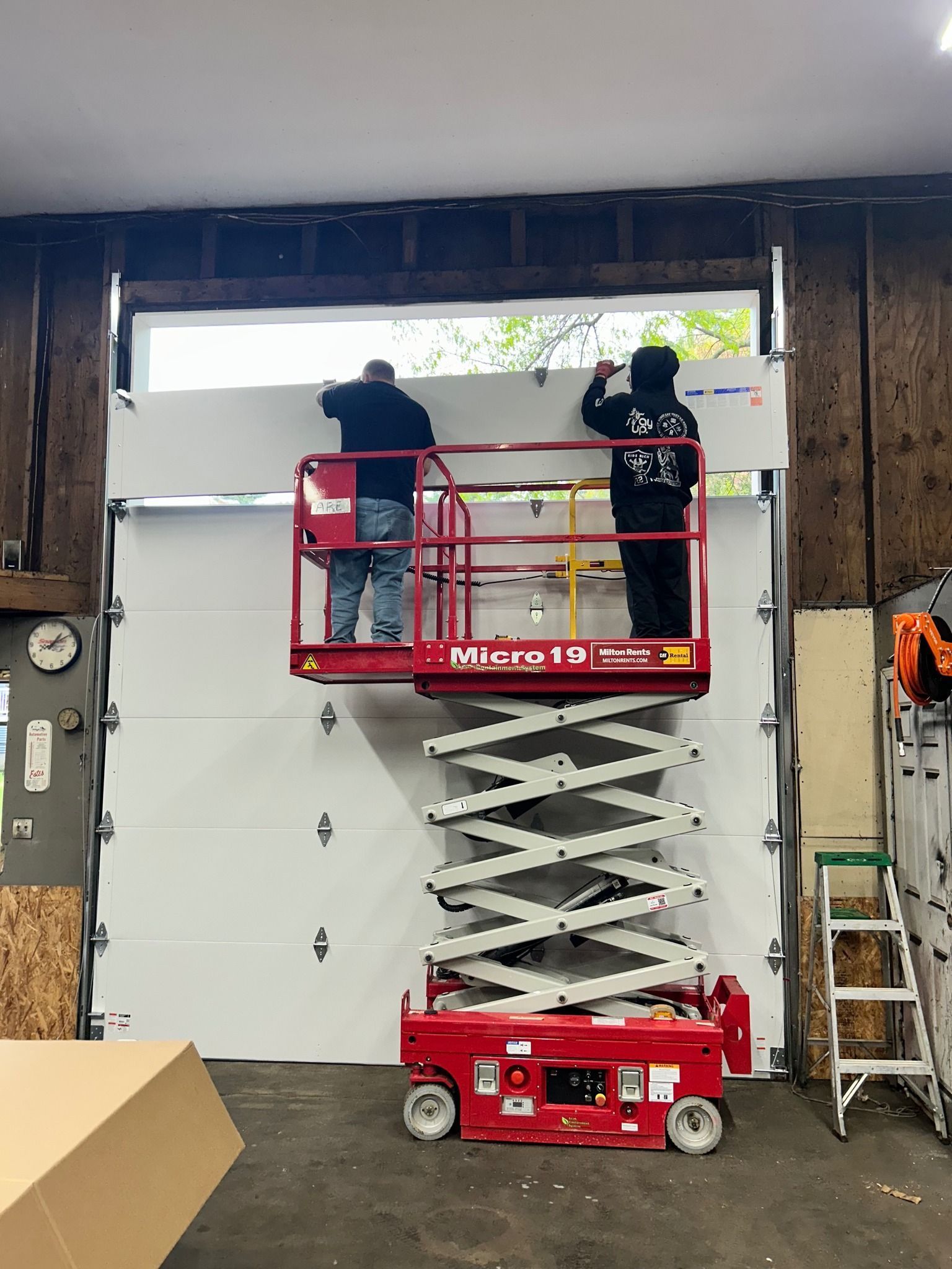 Two people on a red scissor lift install a white garage door panel in a workshop.