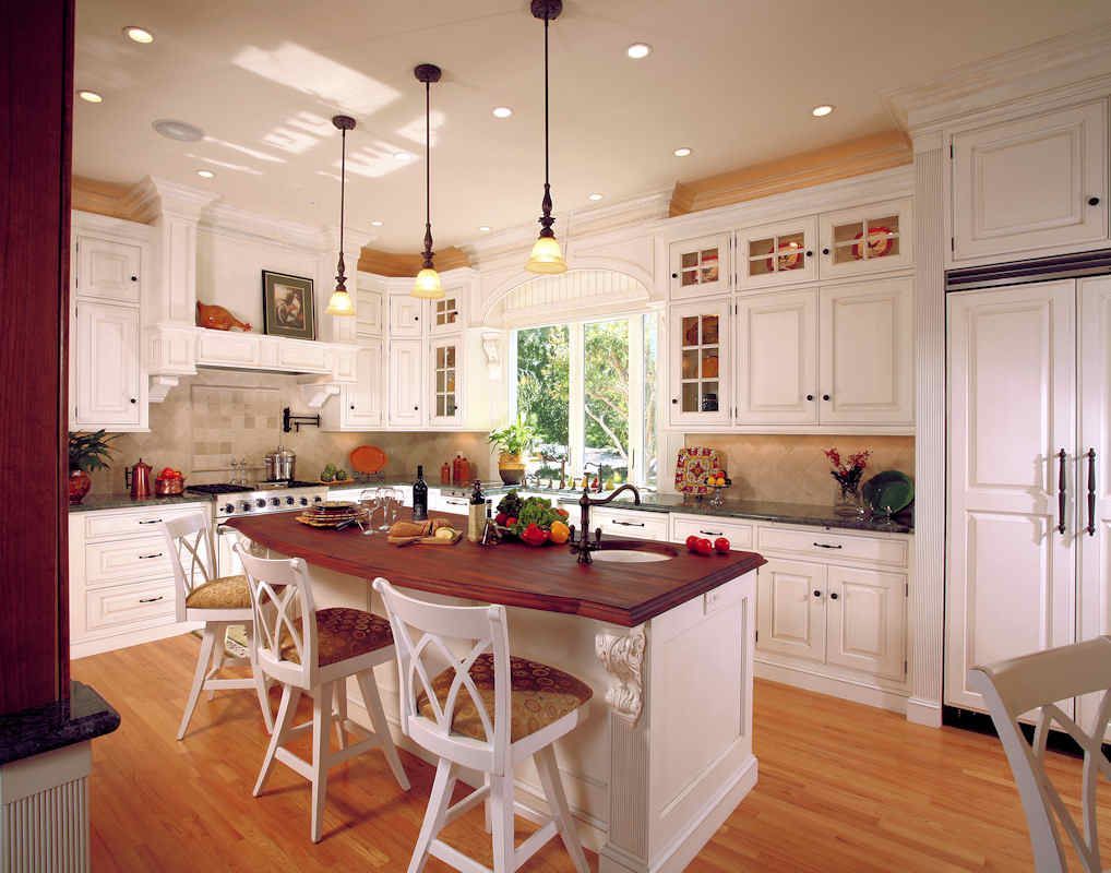 White kitchen with wooden island, countertops, and flooring, lit by pendant lights and natural light from a window.