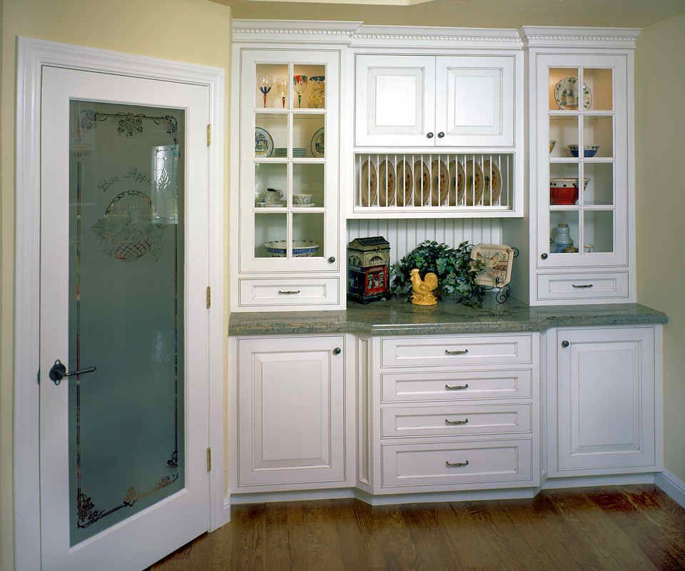 White kitchen cabinets with glass-fronted display cases and a frosted glass pantry door, on wood flooring.