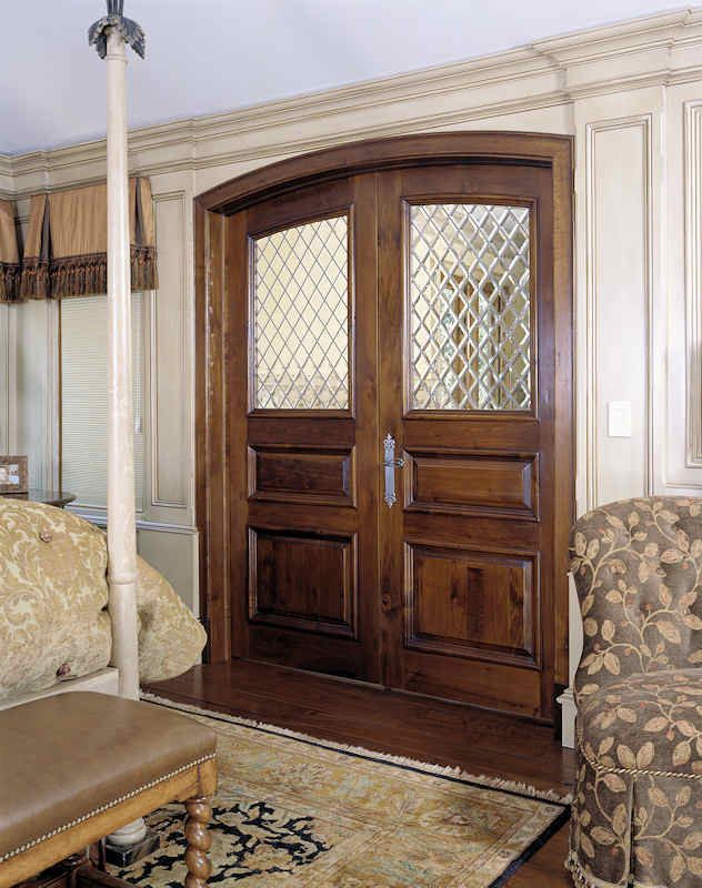 Wooden double doors with diamond-patterned glass panels, in a room with a bed and chair.