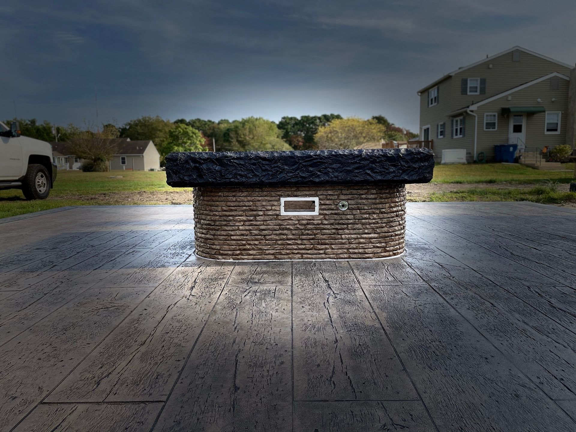 Brick fire pit on a stamped concrete patio, lit by ambient light, with a suburban house in the background.