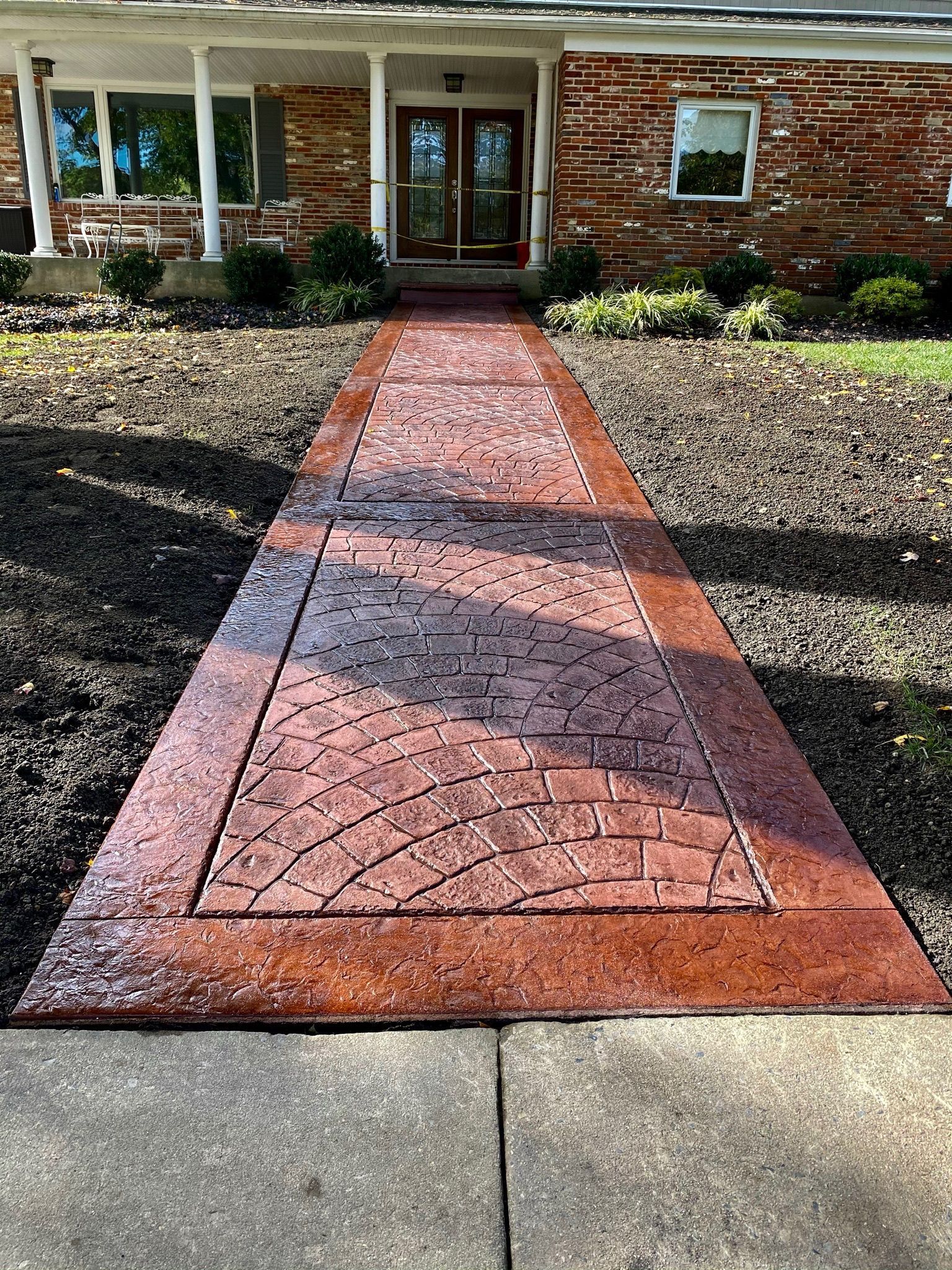 Brick-patterned walkway leading to a brick house with a dark wooden door. Pathway is bordered by a darker brick-colored concrete.