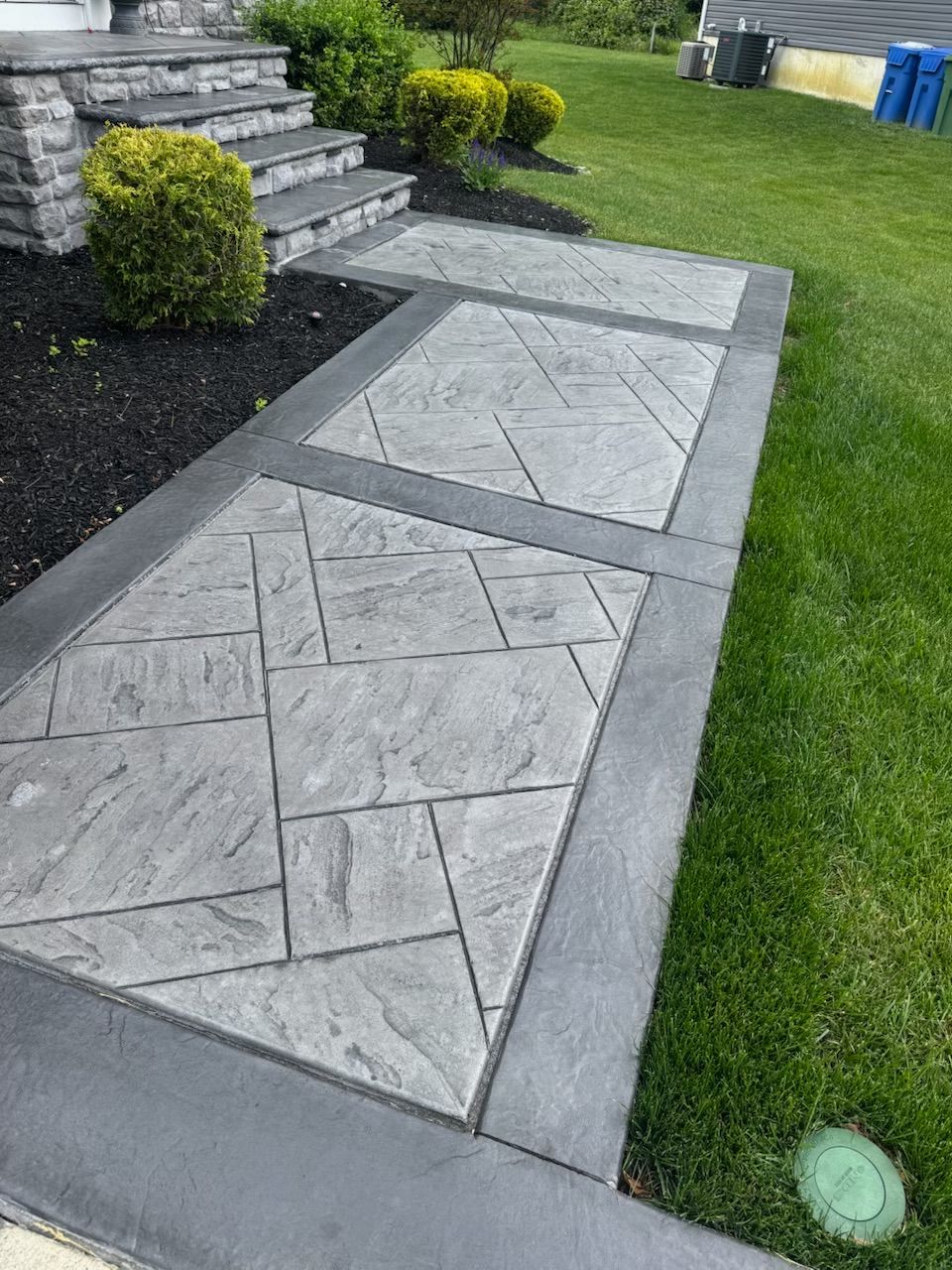 Gray concrete walkway with a patterned design, bordered by a dark gray border, leading to steps and a house entrance.