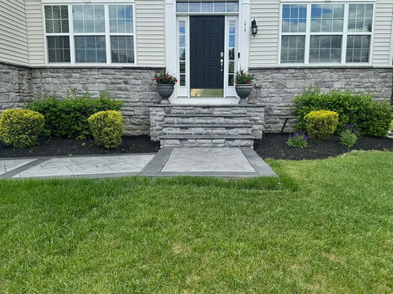 Stone steps leading to a front door with potted plants and symmetrical bushes, flanked by windows.