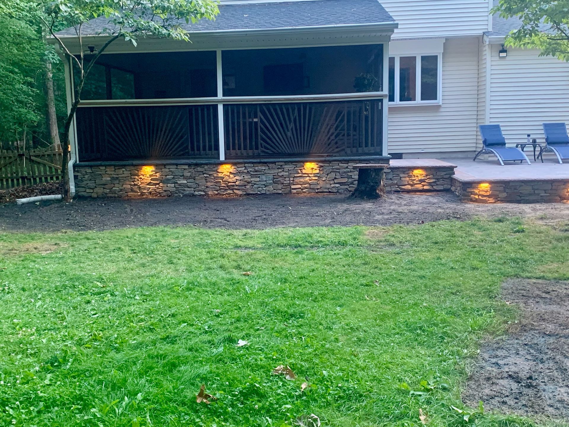 Backyard patio with stone facade, deck, grass, and small retaining walls with lights.
