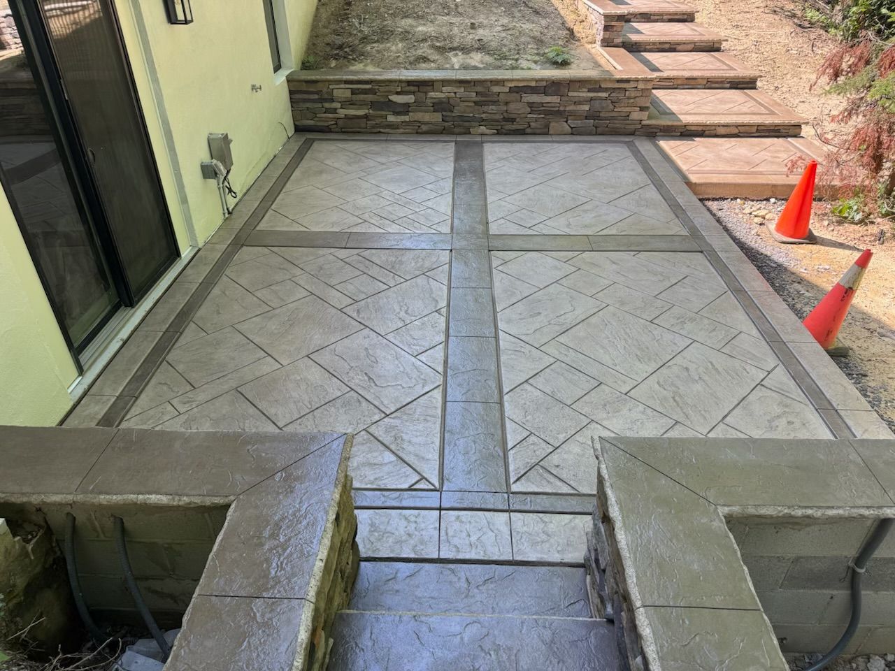 Concrete patio with patterned design, flanked by stone walls and steps, next to a building.