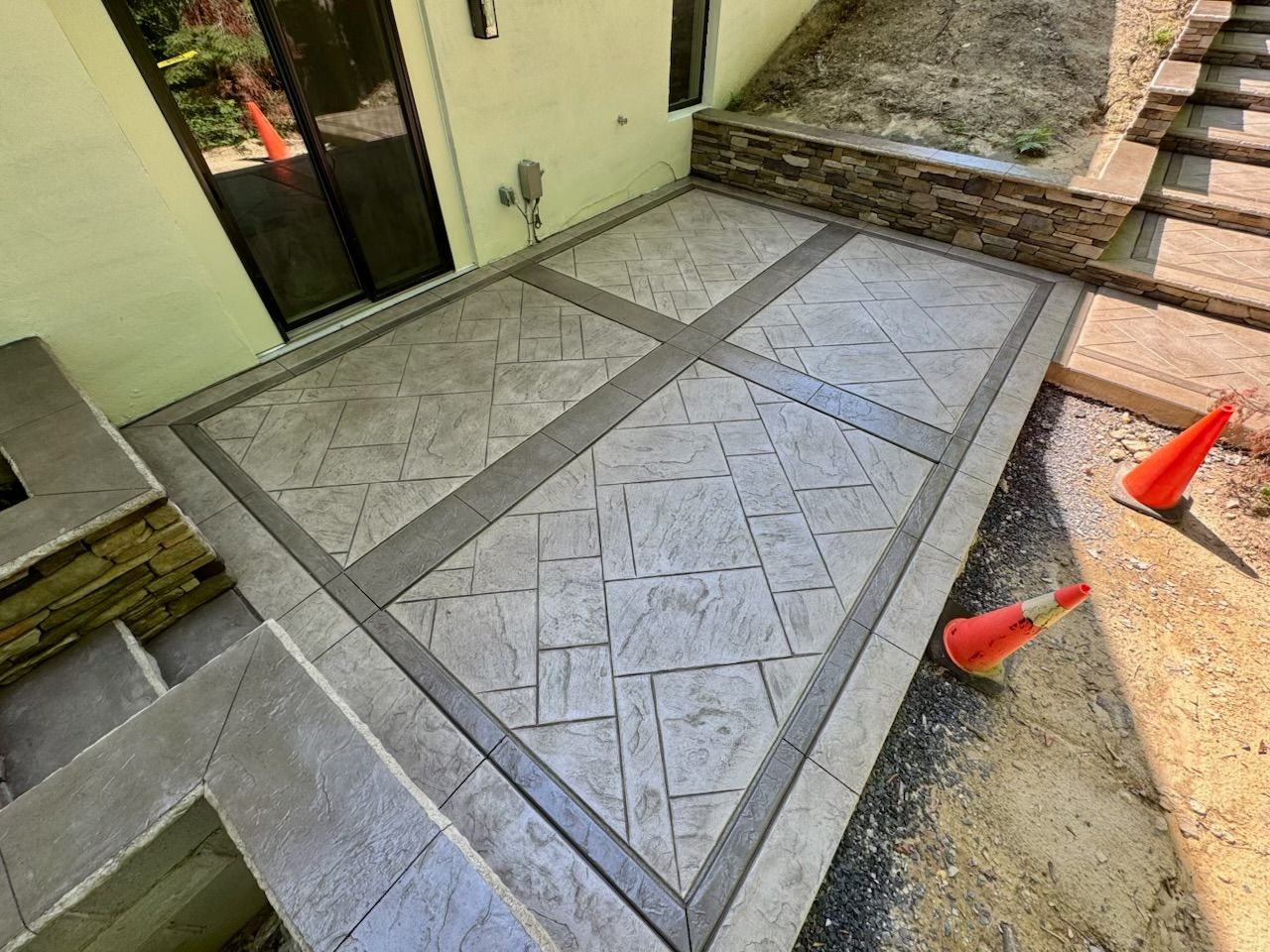 Concrete patio with patterned surface and gray borders, near stairs and building. Orange cones visible.