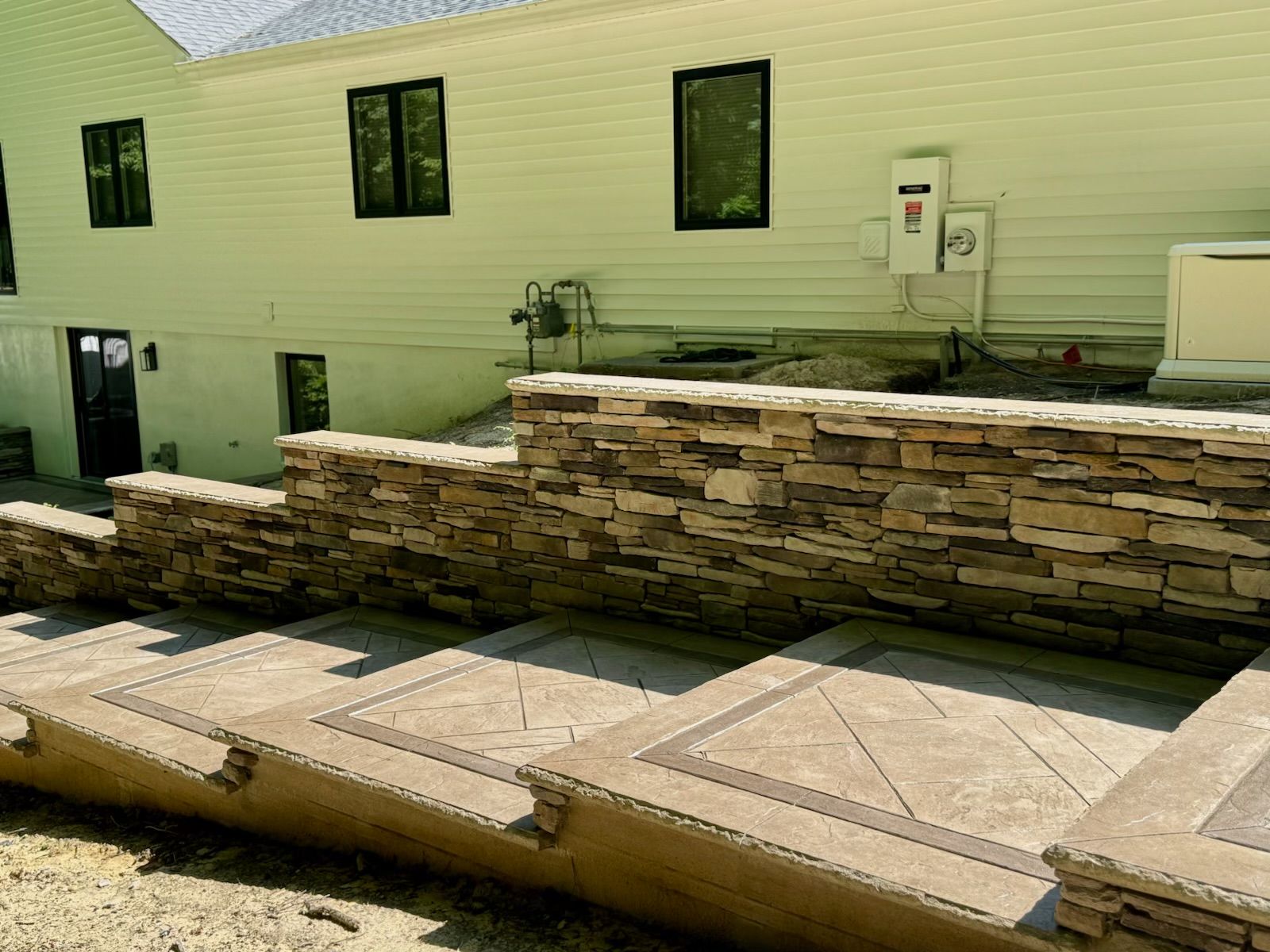 Stone retaining wall with concrete patio in front of a light-colored house with dark window frames.