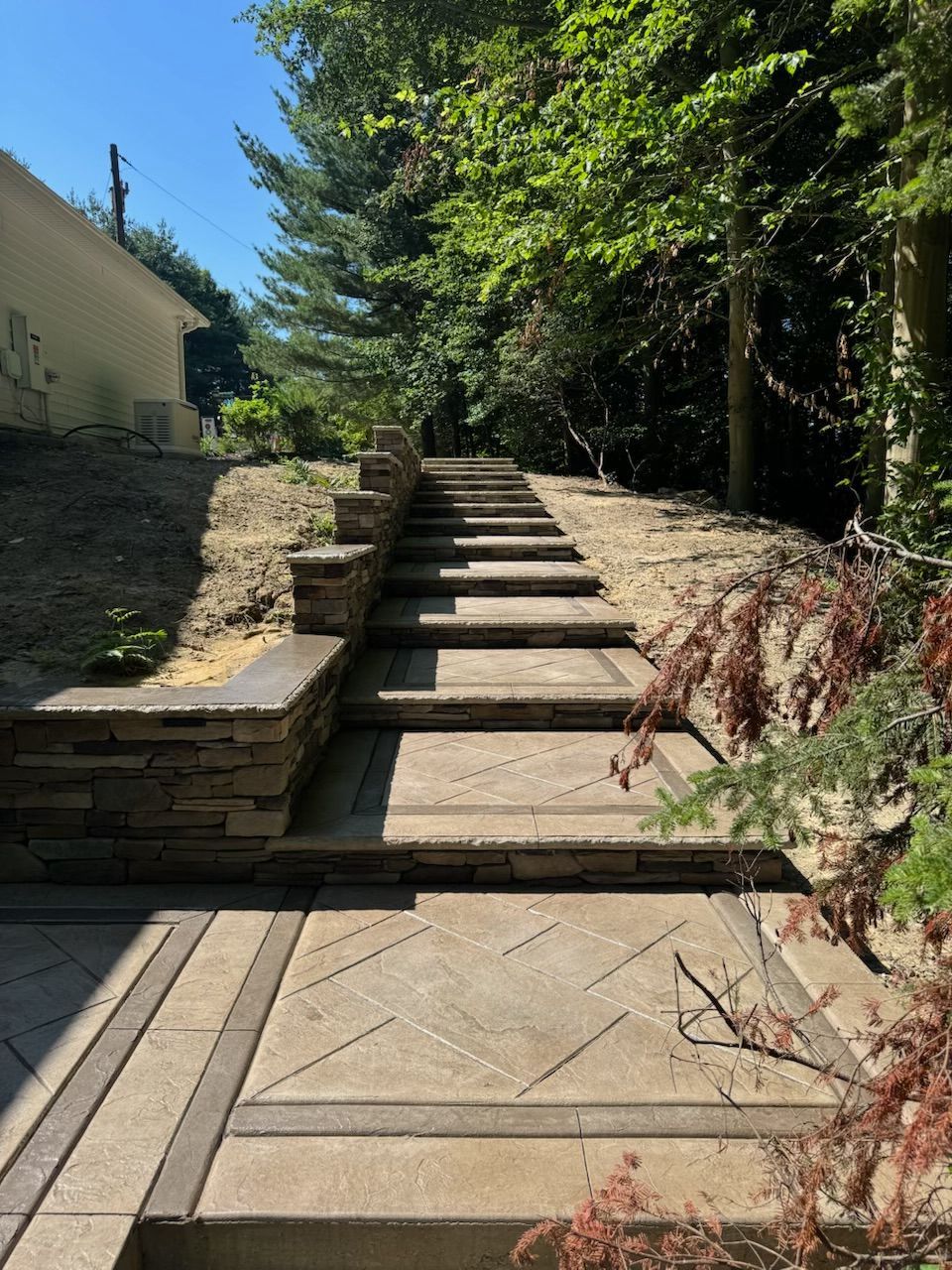 Stone steps leading up a hill with a retaining wall. Trees and a building are in the background on a sunny day.