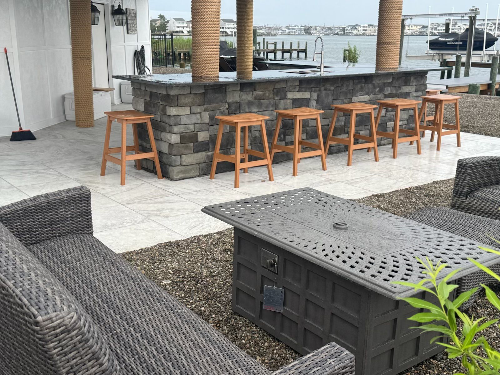 Outdoor bar and seating area overlooking water with brown stools, stone bar, and wicker furniture.