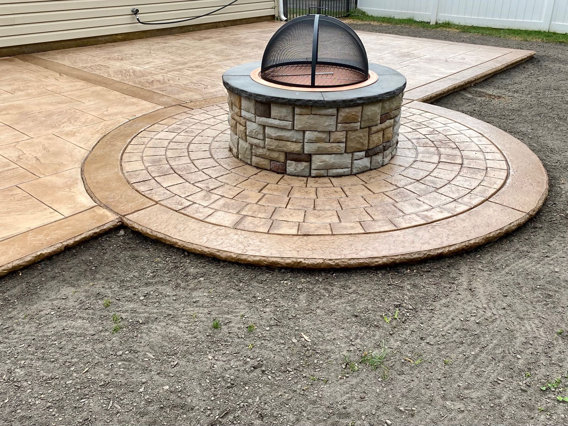 Fire pit on a circular, patterned patio; brown and tan concrete; surrounded by grass.