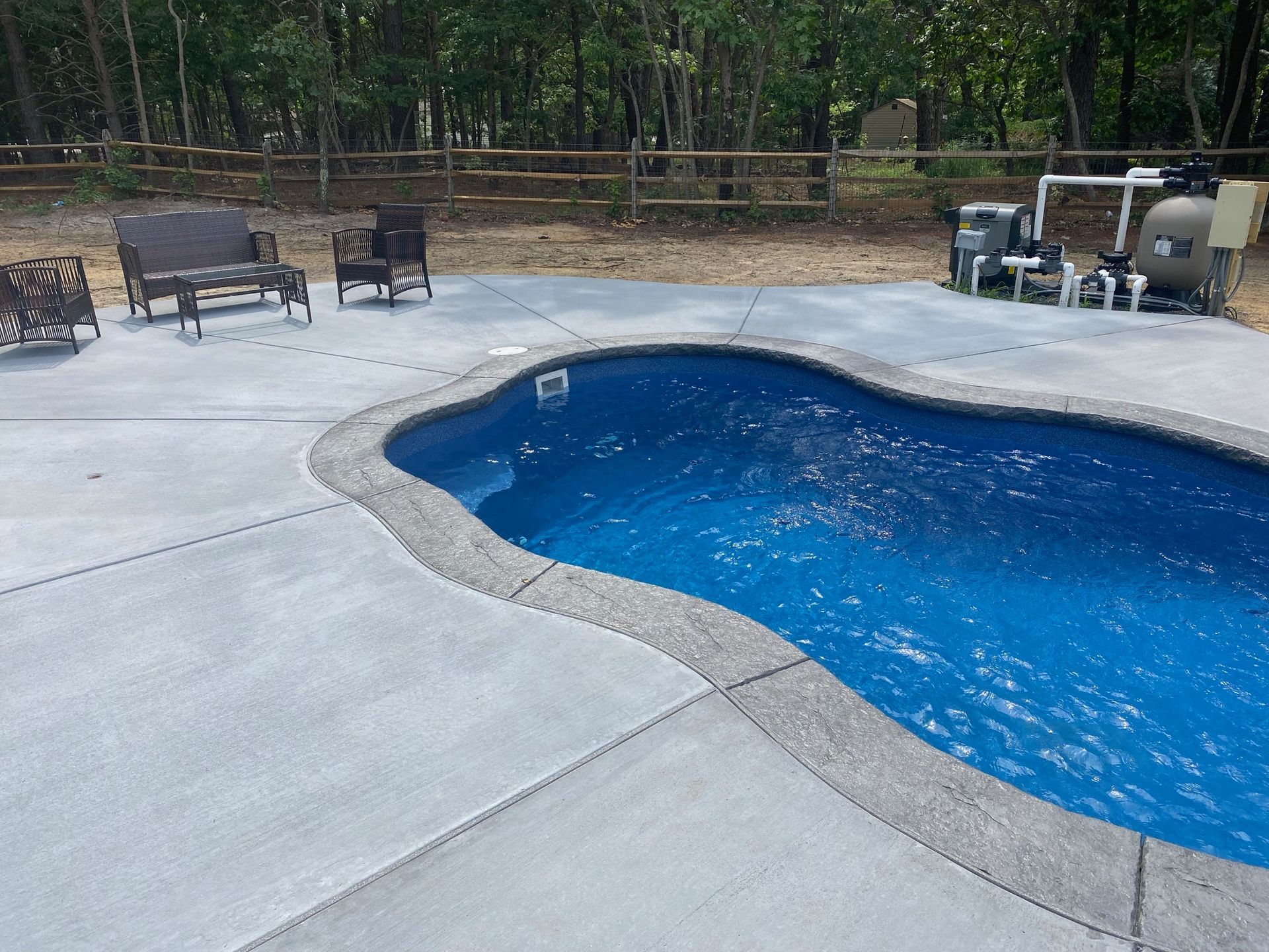 Pool area with blue water, concrete surround, chairs, and equipment in a wooded setting.