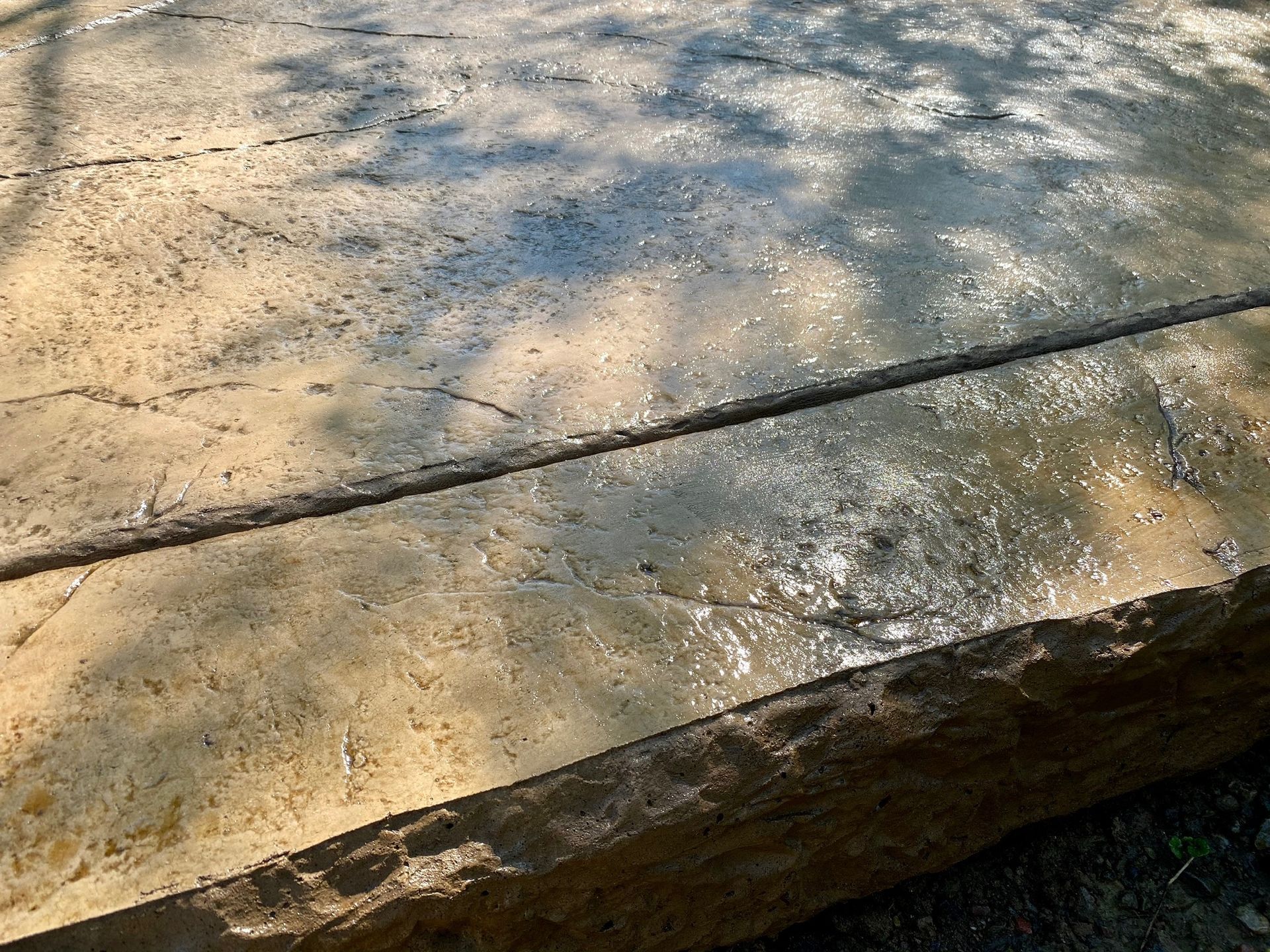 Close-up of wet, light-brown stone slabs. Sunlight reflects off the surface, highlighting cracks and rough edges.