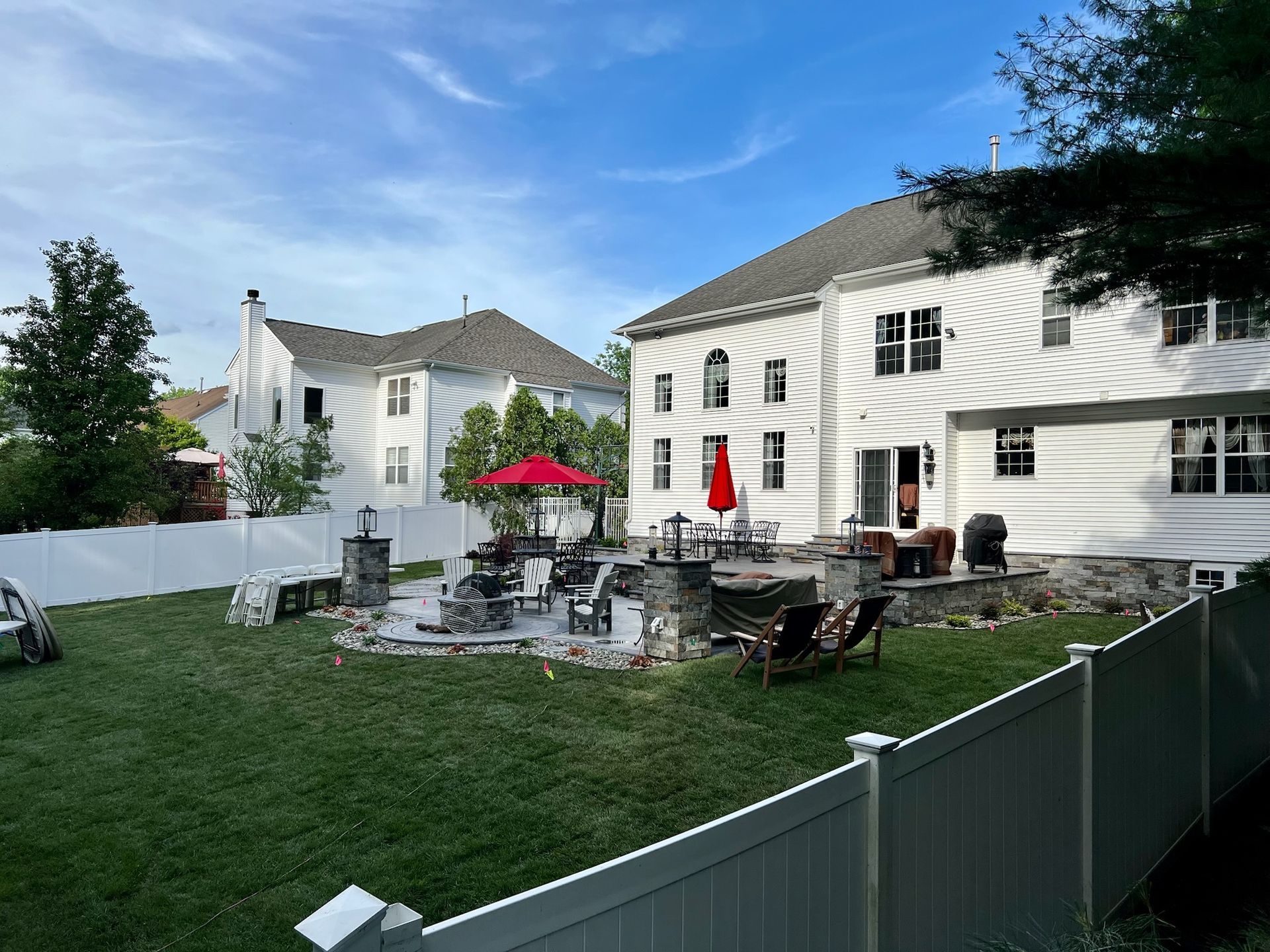 Backyard with white house, patio, red umbrella, lawn, and white fence. Blue sky.