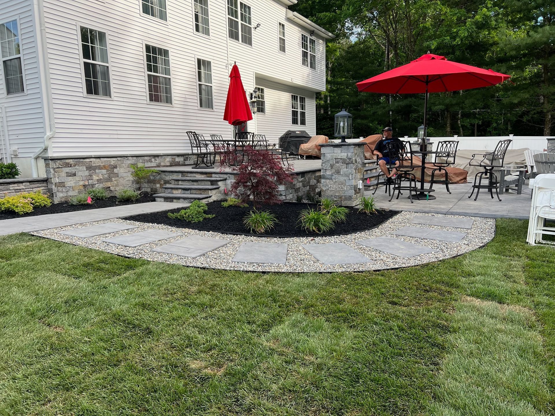 Backyard patio with seating, red umbrellas, stone path, and manicured lawn.