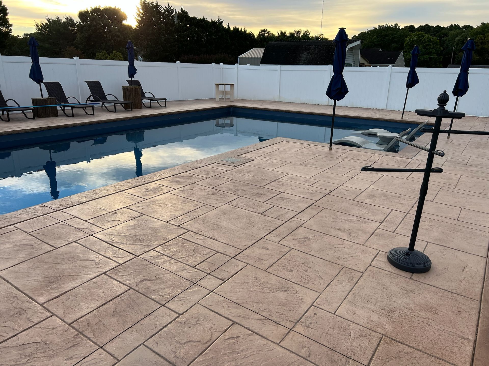 A pool deck with a rectangular pool and lounge chairs, surrounded by a white fence and umbrellas.