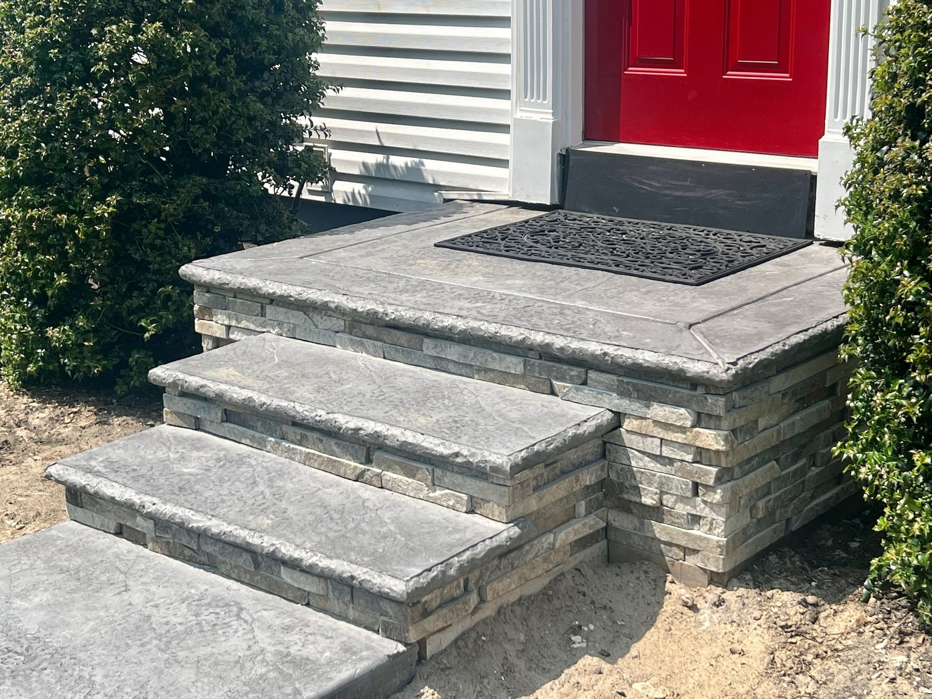 Stone steps leading to a red door. Stone veneer on the sides with greenery and a welcome mat.