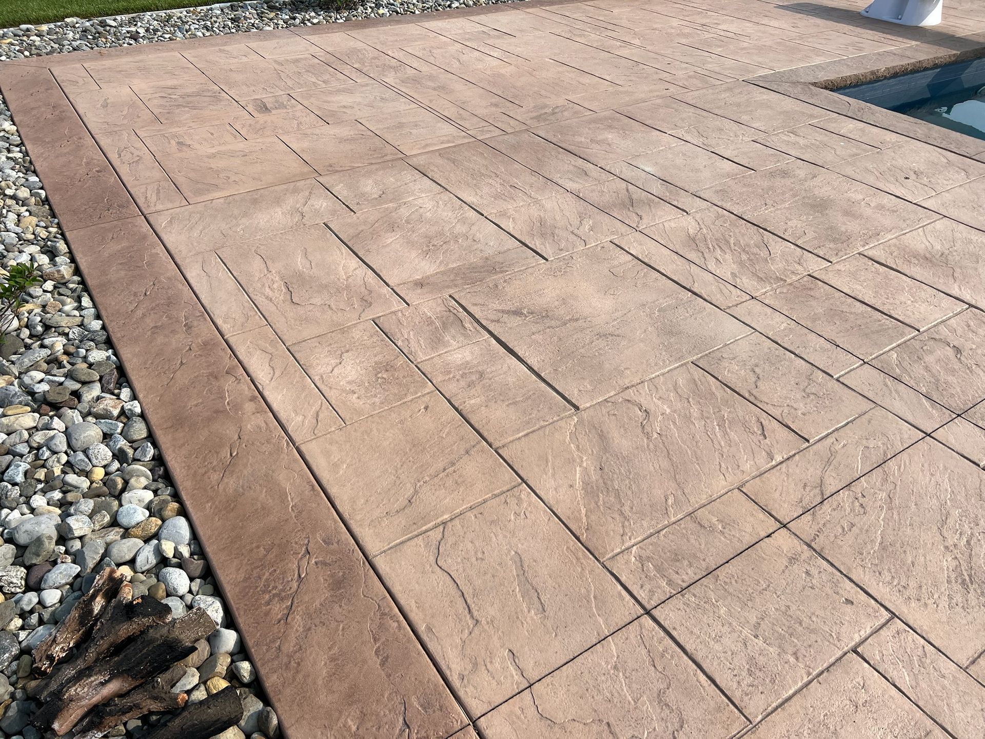 Tan stamped concrete patio with a brown border next to a pool and gravel landscaping.
