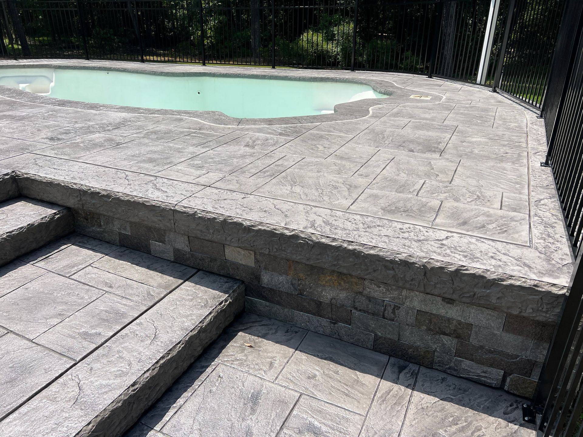 A pool deck with steps and a retaining wall with a decorative brick pattern, enclosed by a black metal fence.