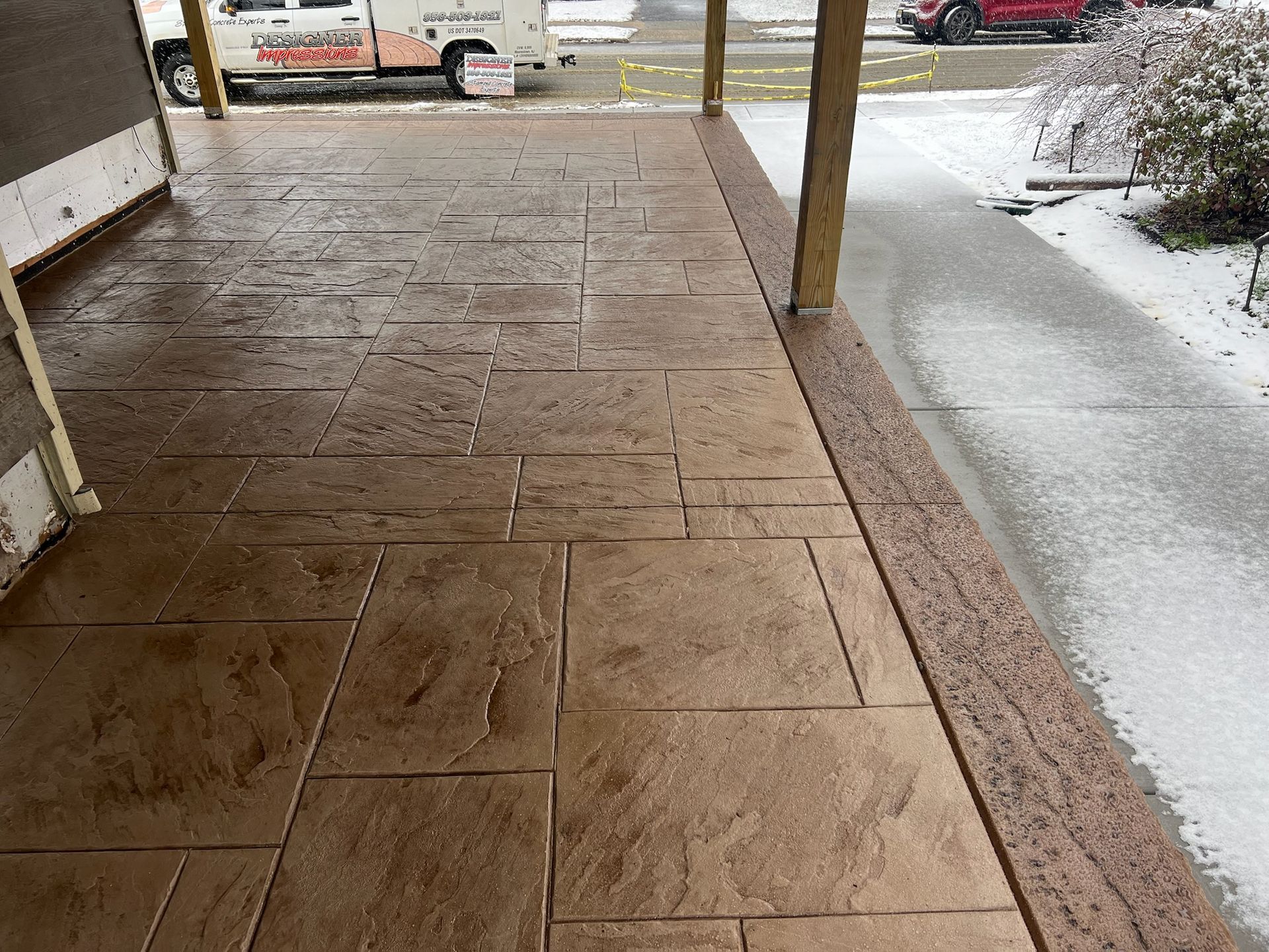 Brown stamped concrete porch with snow, next to a snowy sidewalk.