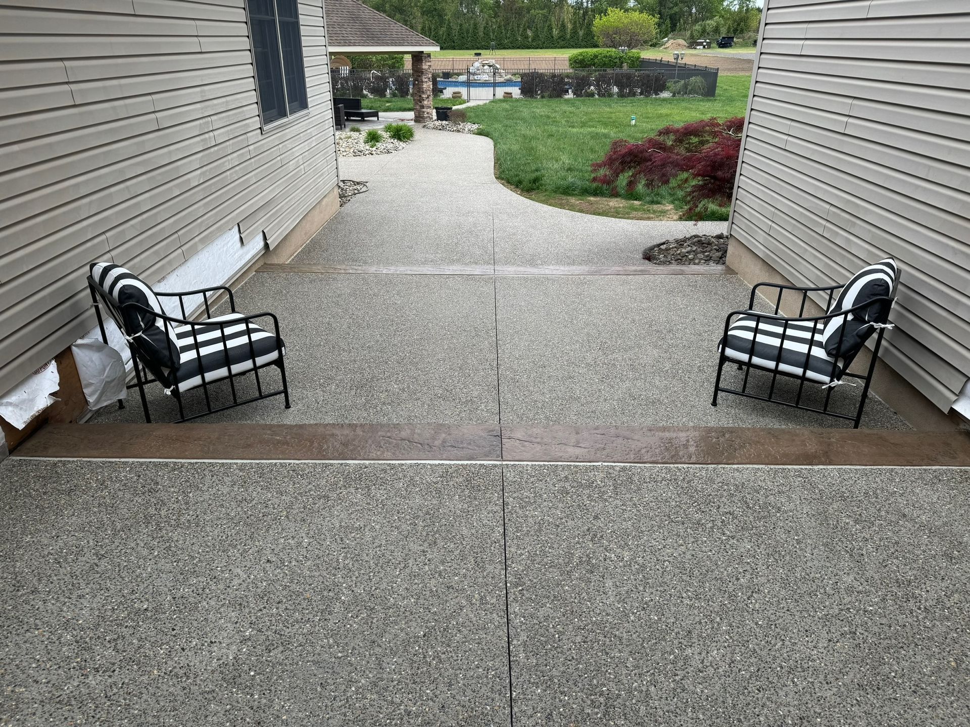 Two striped chairs on a concrete patio between two houses; grassy backyard with a pool is visible.