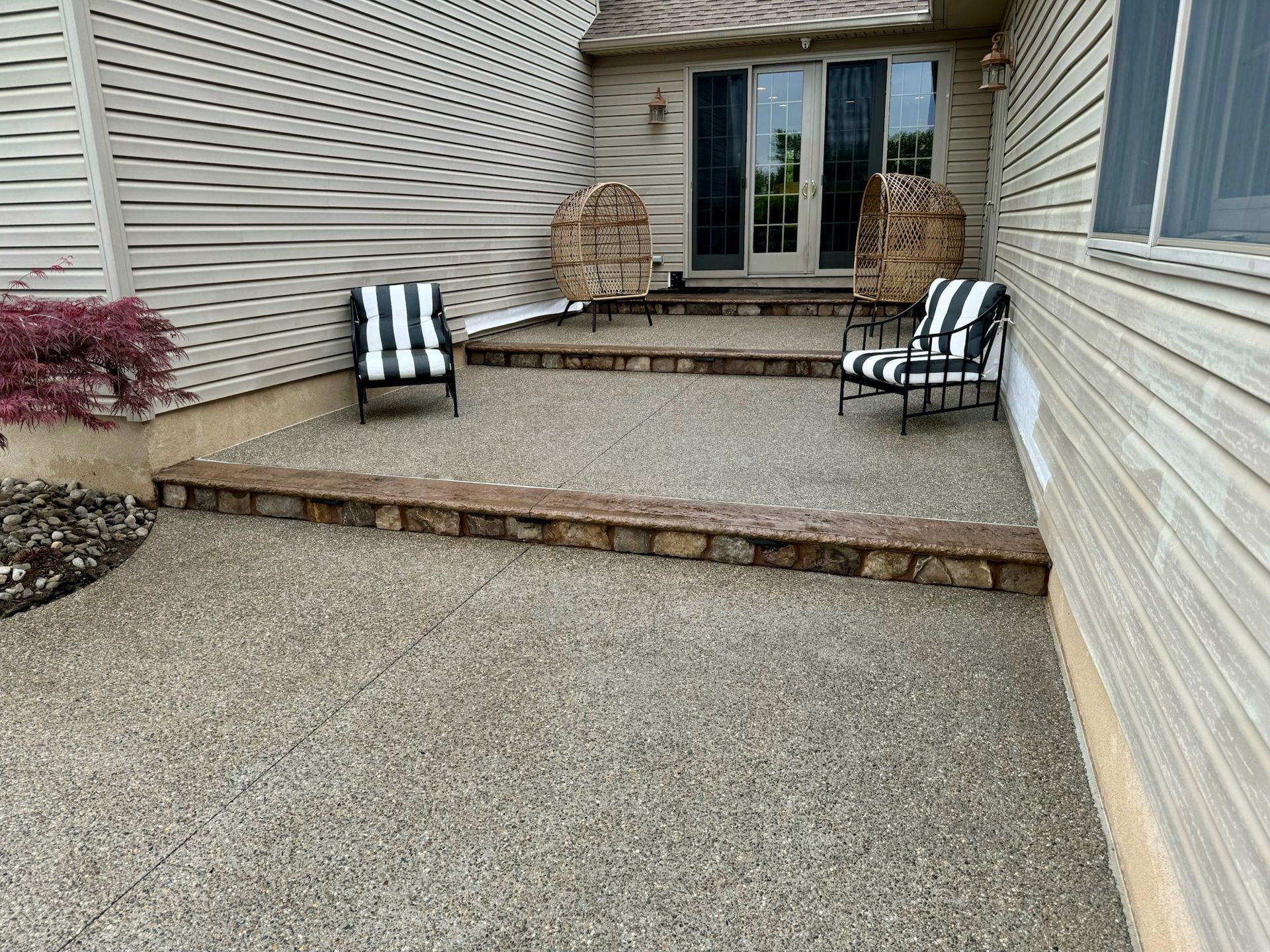 Two-tiered concrete patio with steps, two striped chairs, and a door to a house.