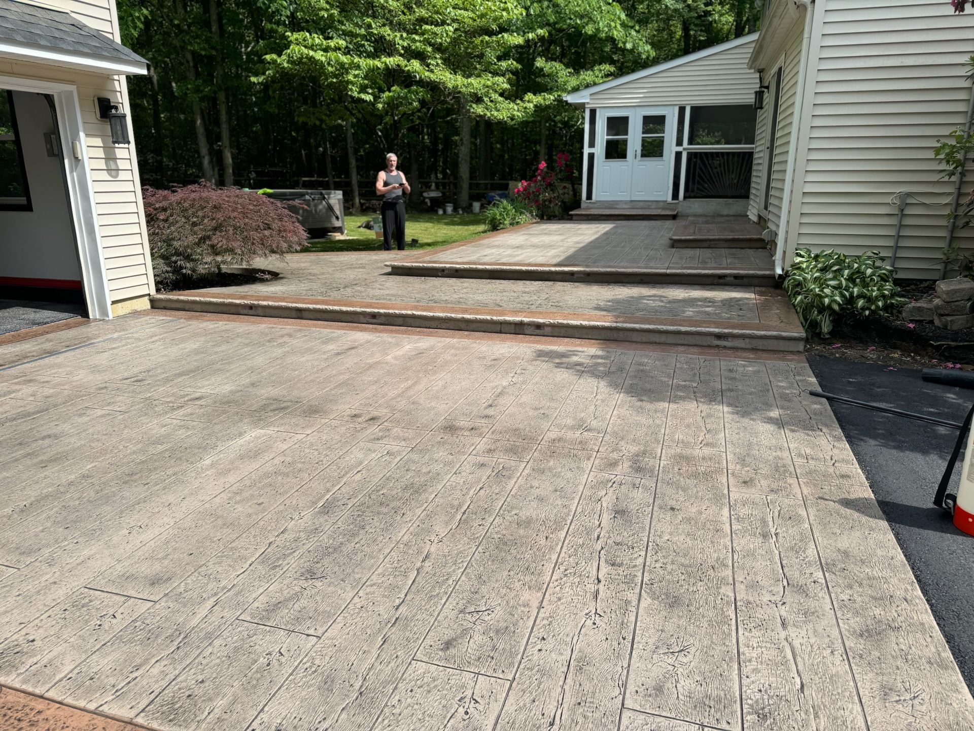 Person standing on a paver patio next to a house with a covered entrance, a driveway, and landscaping.