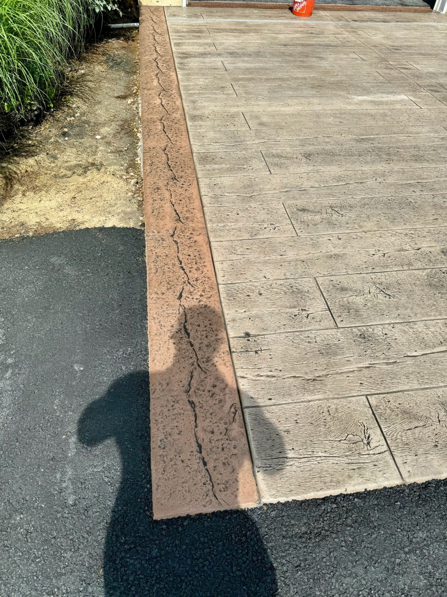 Shadow of person on asphalt next to concrete patio with brown border.