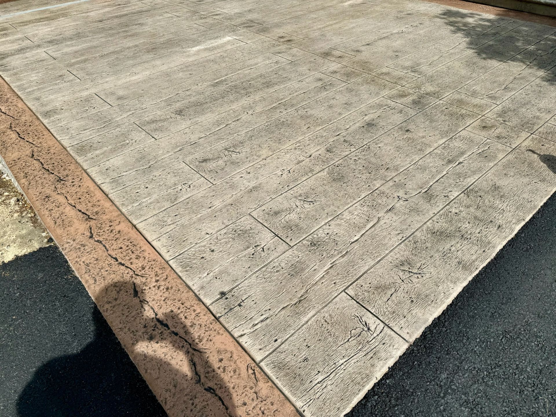 Concrete patio with stamped brick pattern, bordered by a textured brown edge and black asphalt.