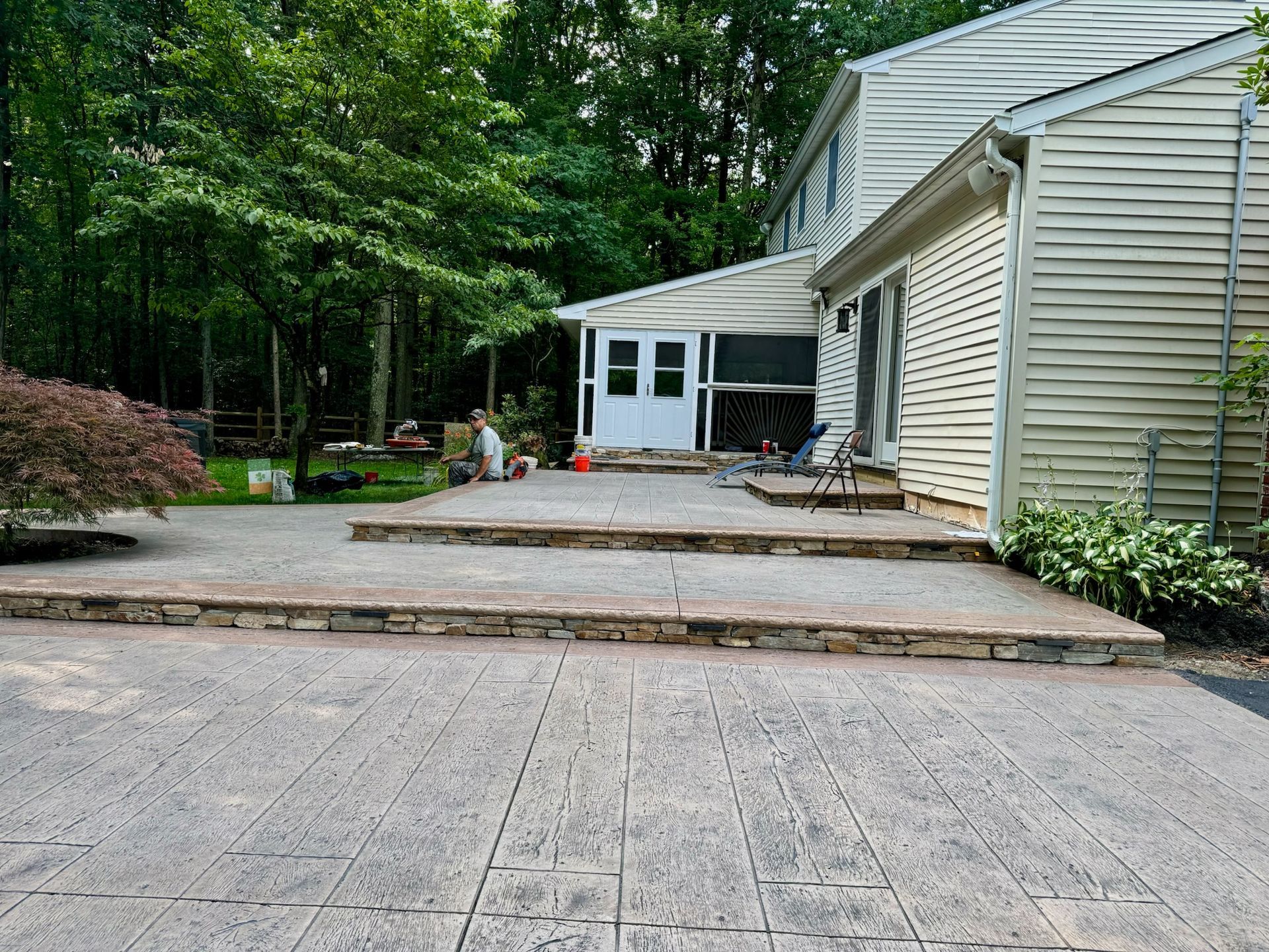 A two-story house with a patio and steps; trees in the background; patio is gray with textured pattern.