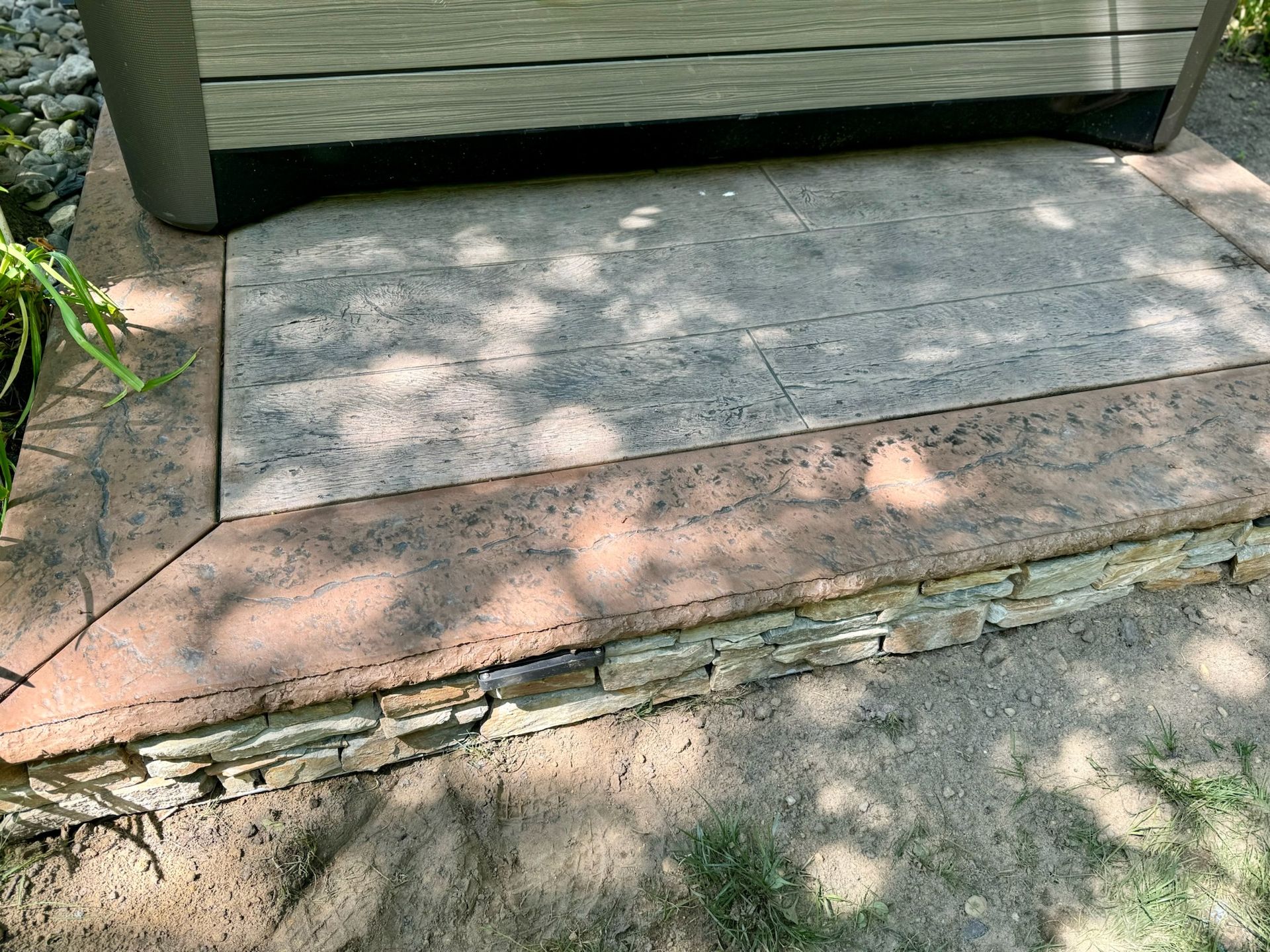 A hot tub rests on a stone patio with a stone border, surrounded by earth.