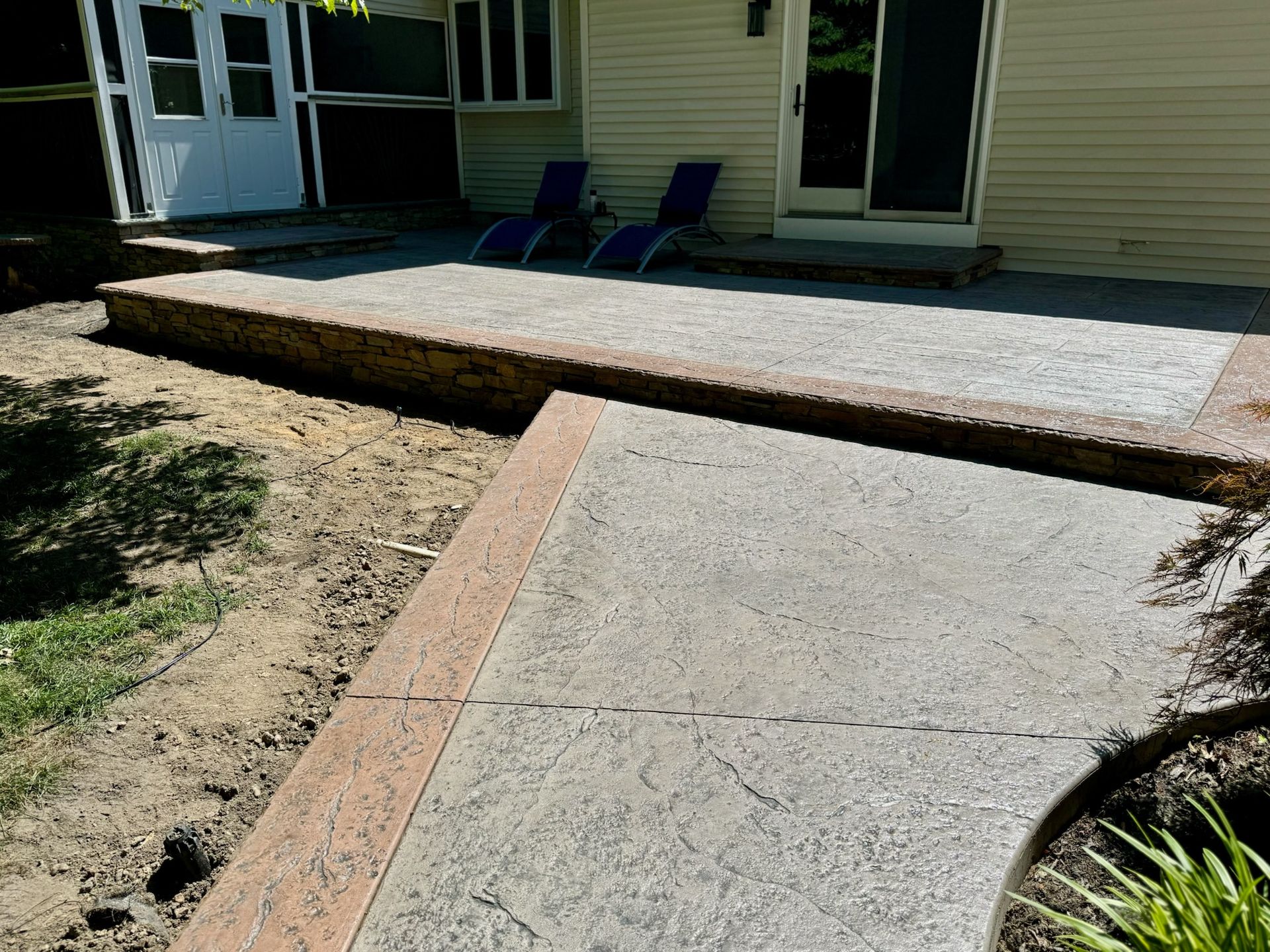 Concrete patio with a raised border and walkway leading to it. Beige siding, two blue chairs.