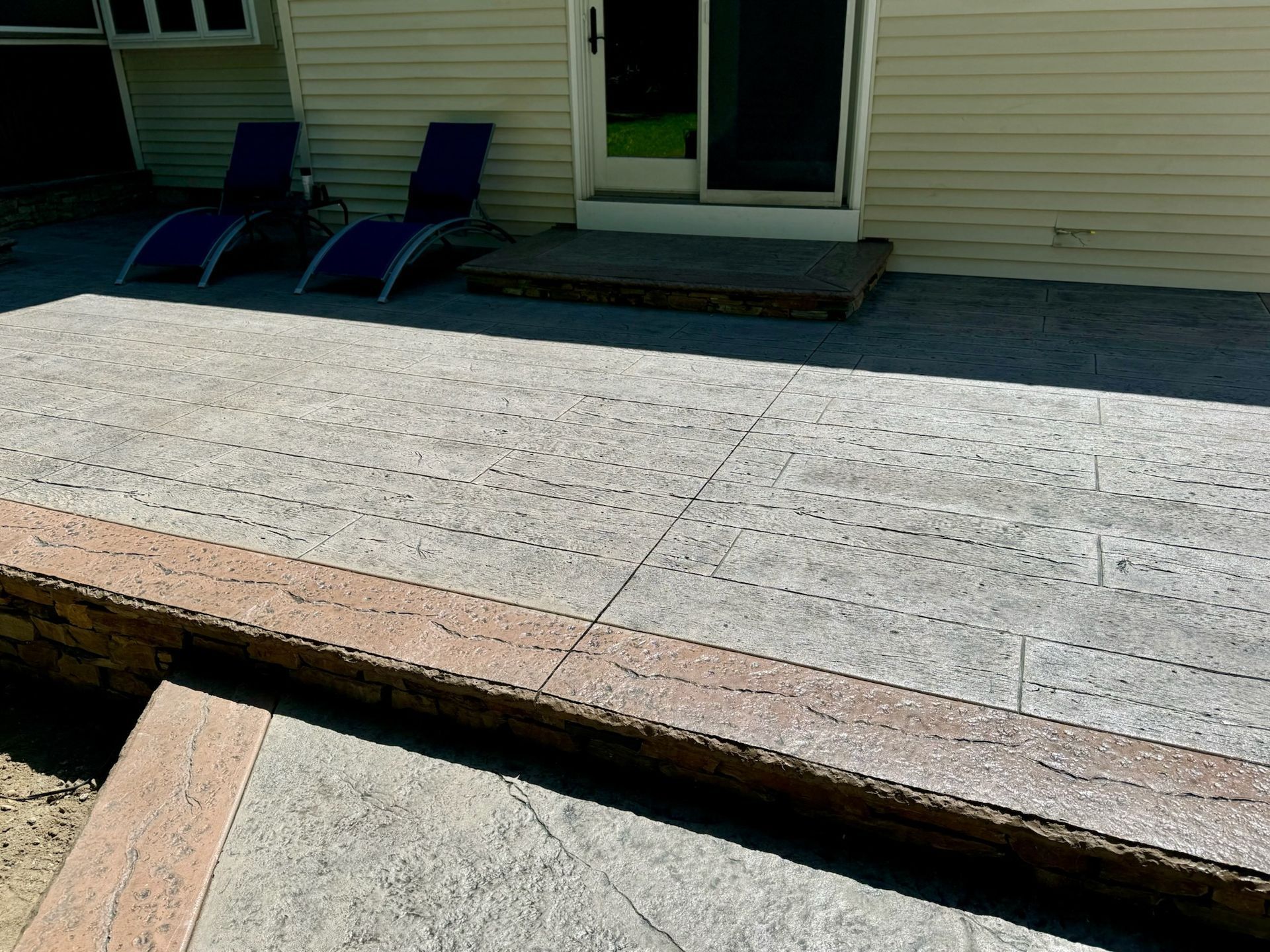Concrete patio with two lounge chairs, steps to sliding door of a beige house.