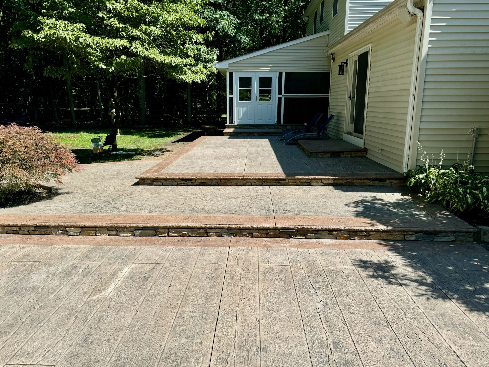 Concrete patio with steps, bordered by reddish bricks, next to a house with a sunroom.
