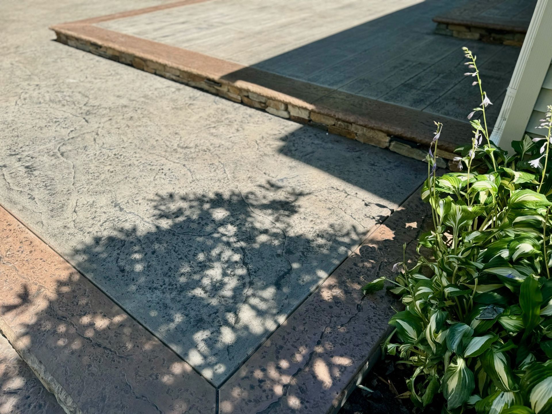 Concrete patio with textured surface and colored border, next to plants and house.