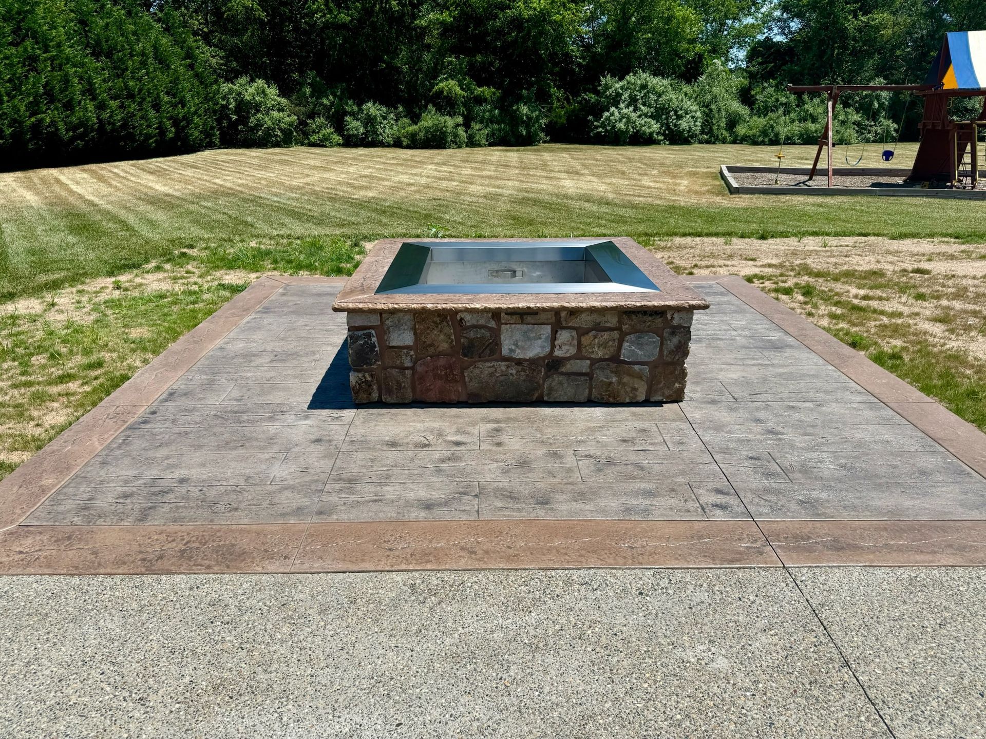 Fire pit with stone base and concrete patio, in a park setting.