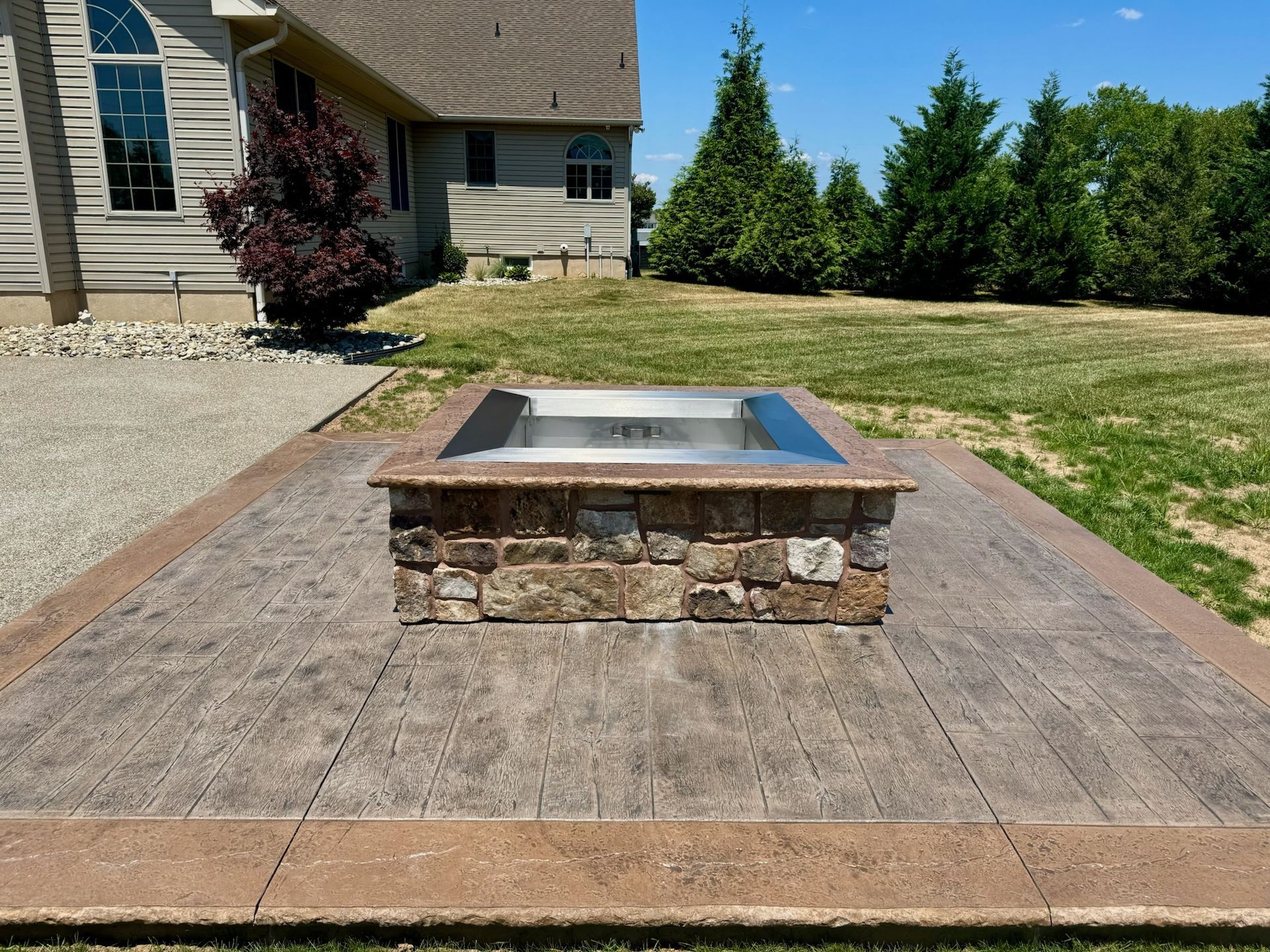 A stone fire pit sits on a concrete patio in a backyard, with a house in the background.