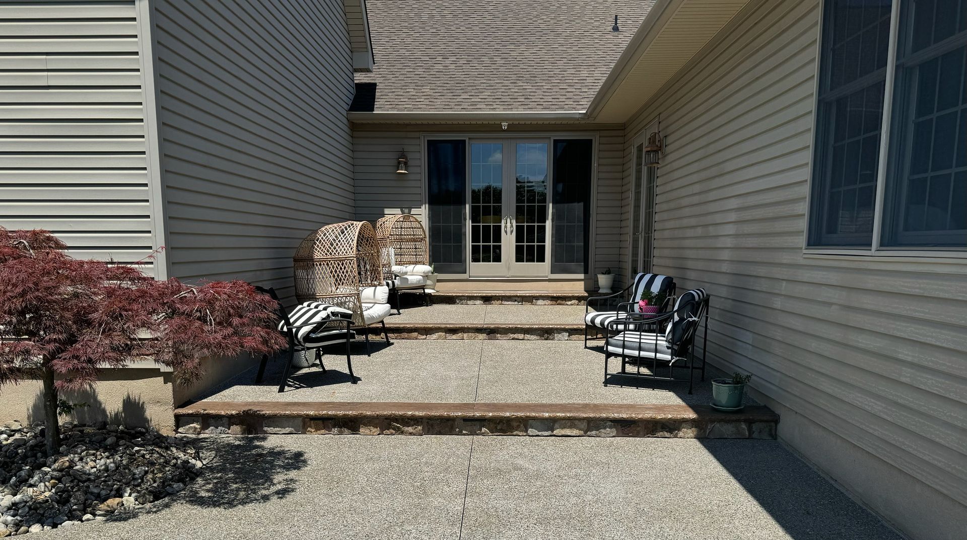 Patio with gravel, steps, chairs, and double glass doors. Home exterior with light siding and a red-leaved shrub.