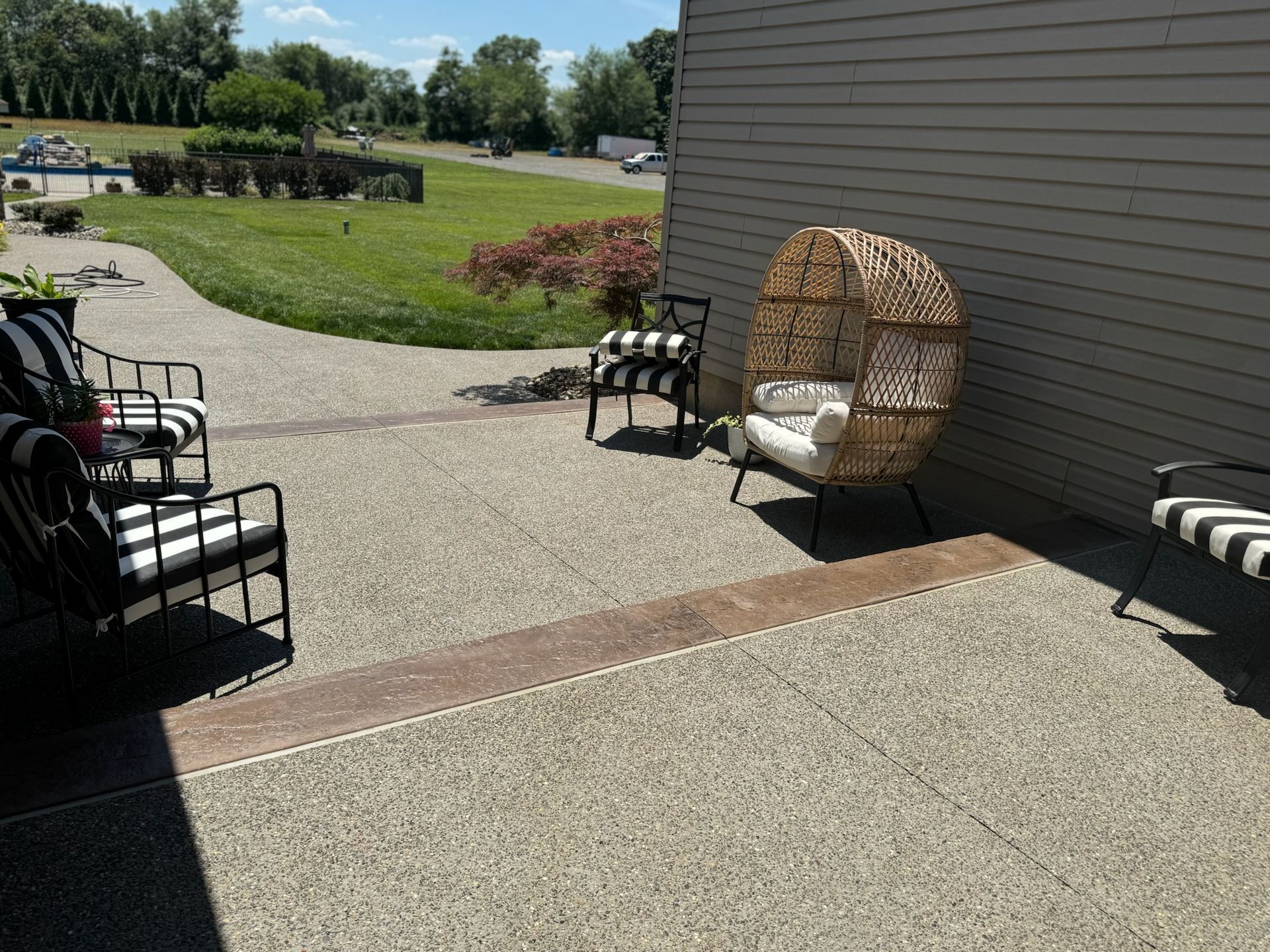 Outdoor patio with seating: black and white striped chairs, wicker egg chair, concrete surface, grassy yard.