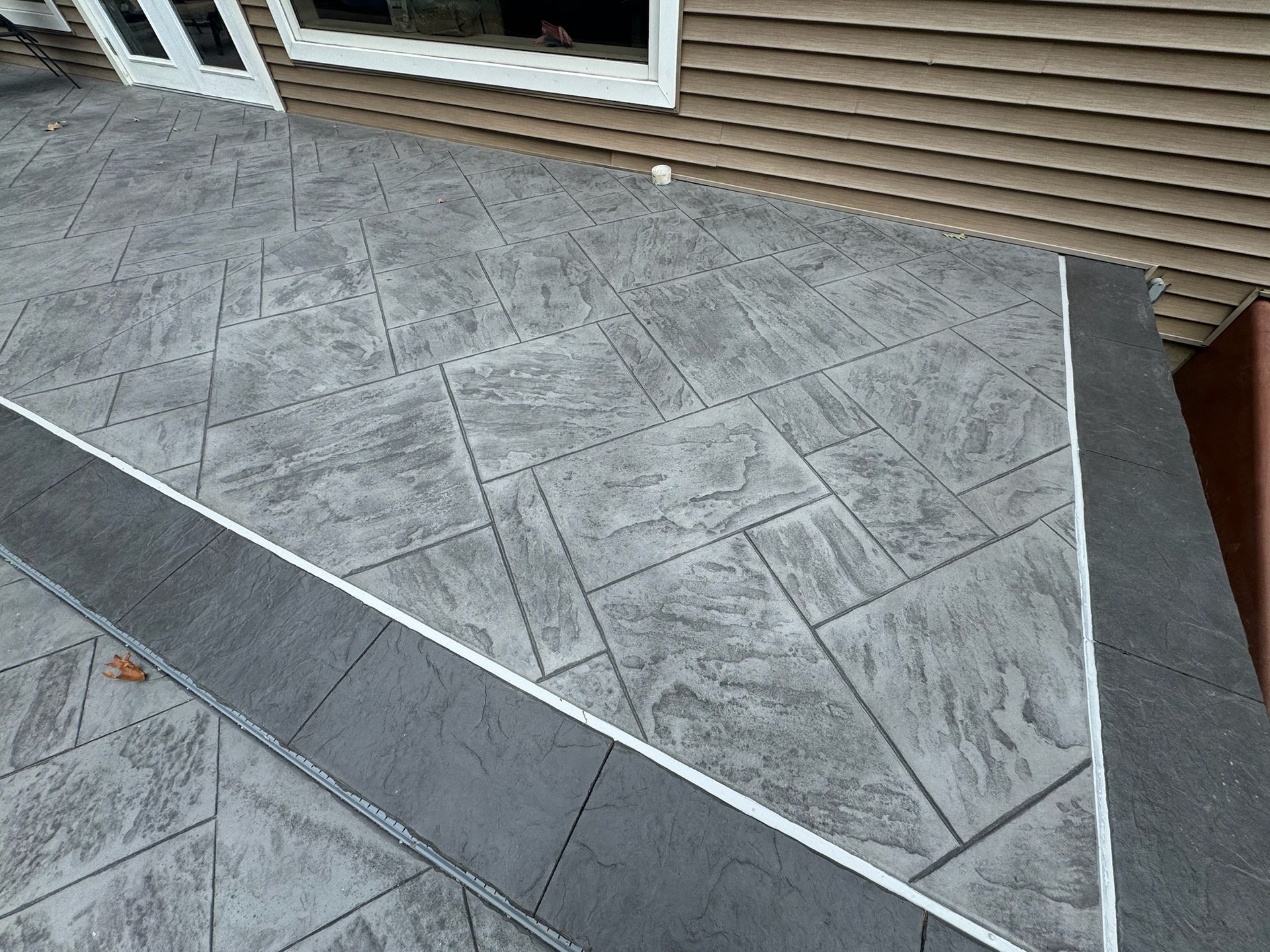 Gray stamped concrete patio with dark border and white trim against a brown house.