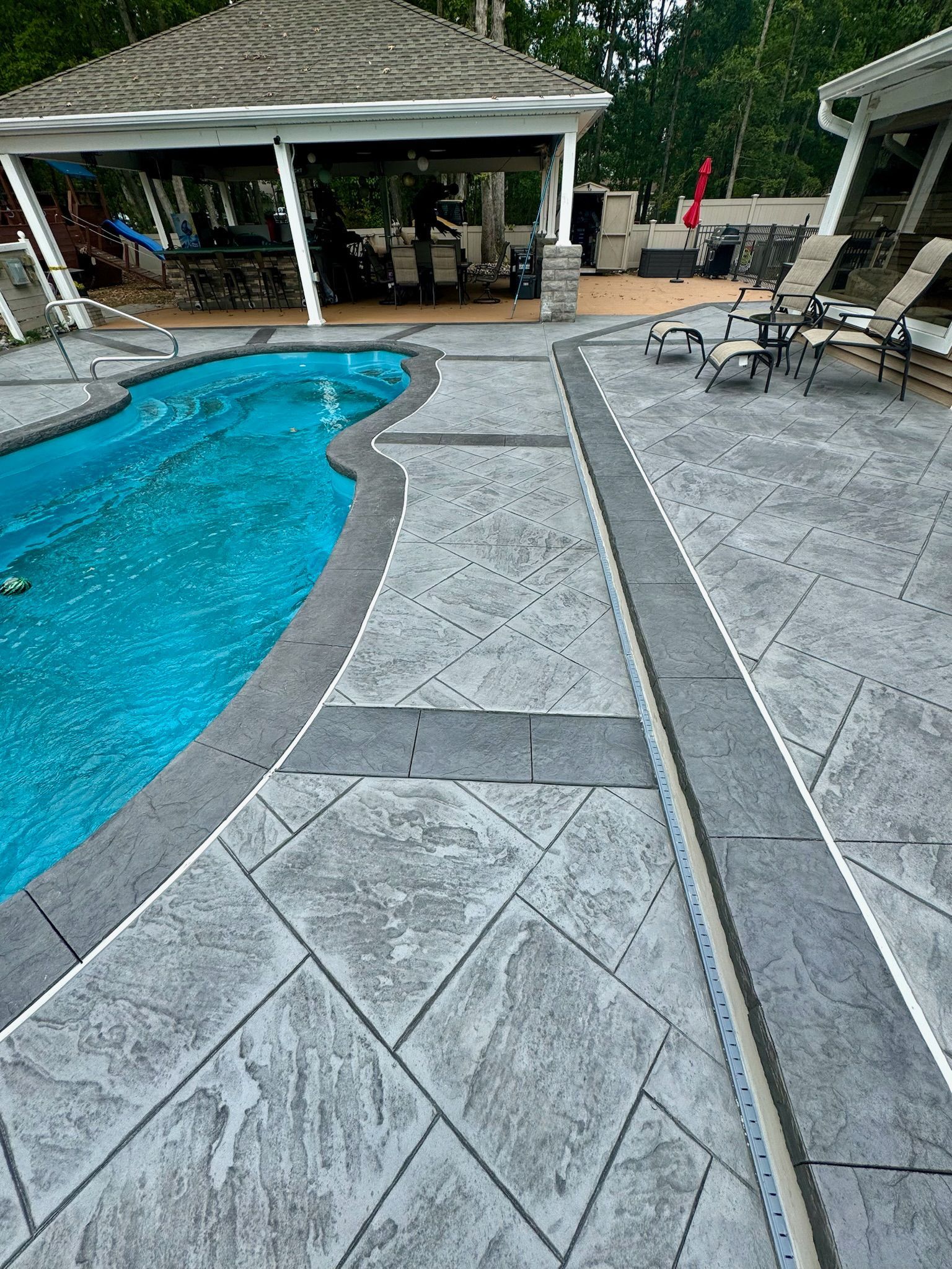 Poolside stamped concrete patio with pergola, pool, and lounge chairs; gray tones.