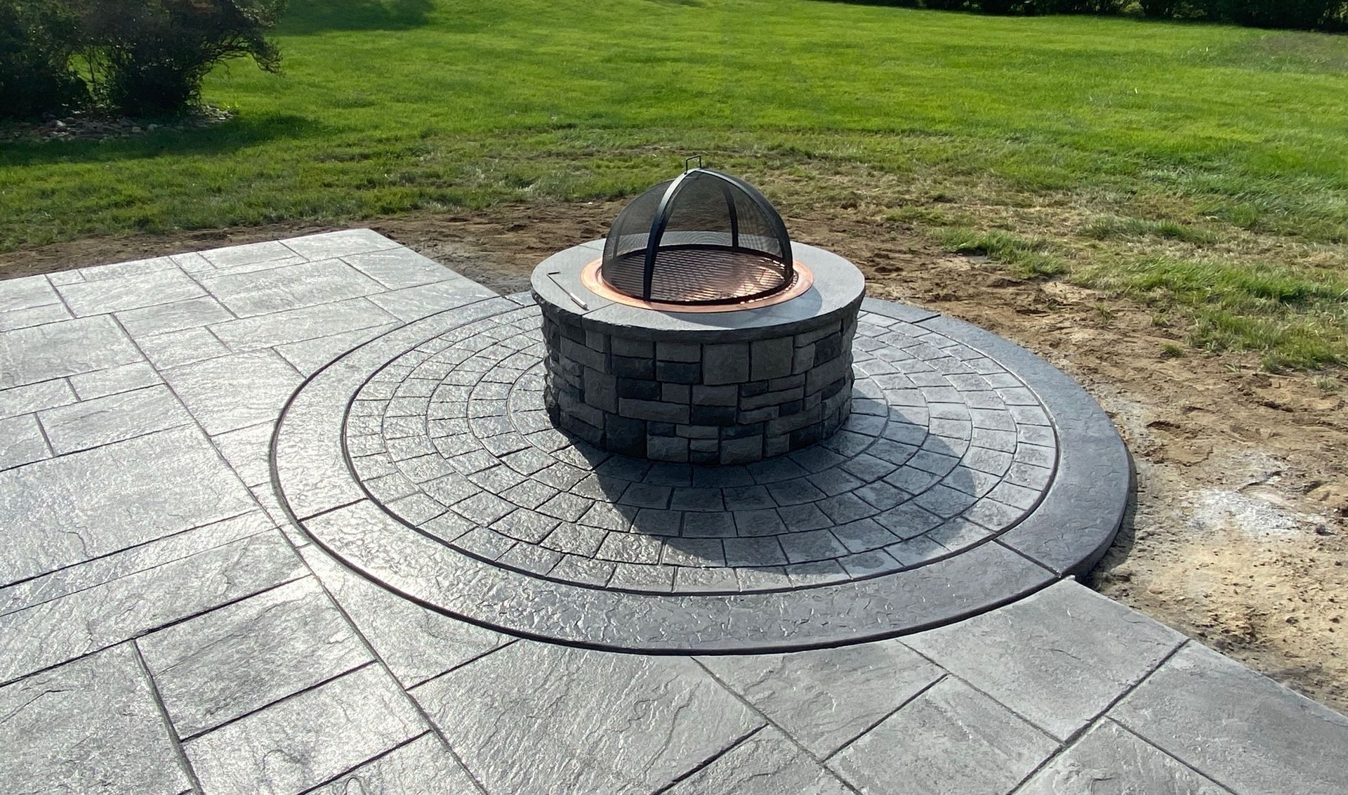 Fire pit on a circular patio with concrete pavers, surrounded by grass.